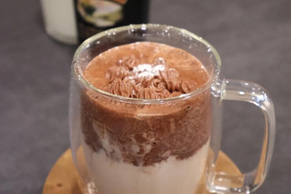 Baileys hot chocolate in a clear mug with whipped cream and cocoa powder topping, layered with cream.