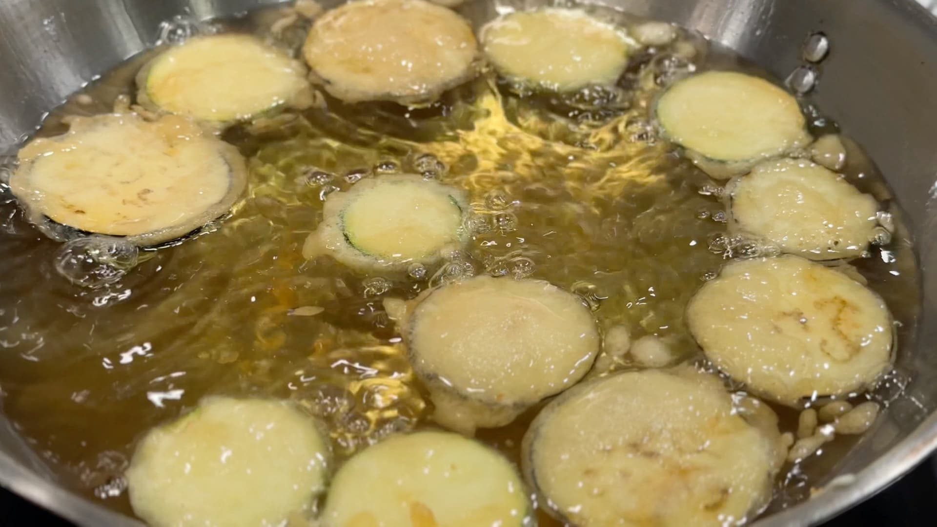 Sliced vegetables frying in golden oil in a stainless steel pot, including zucchini, eggplant, and yam turning golden brown.