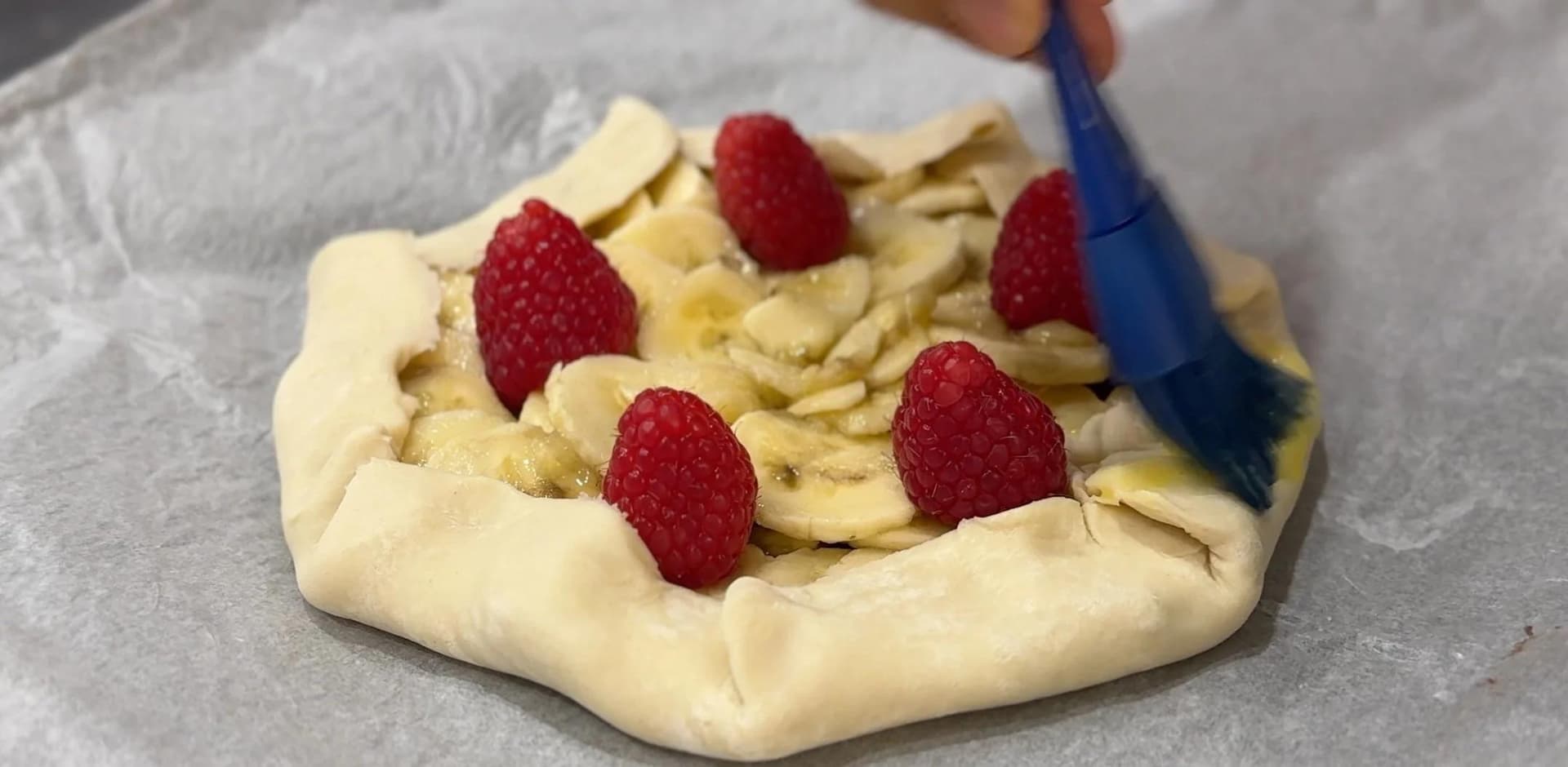 Banana Nutella galette with pleated edges topped with fresh raspberries, being brushed with egg wash.
