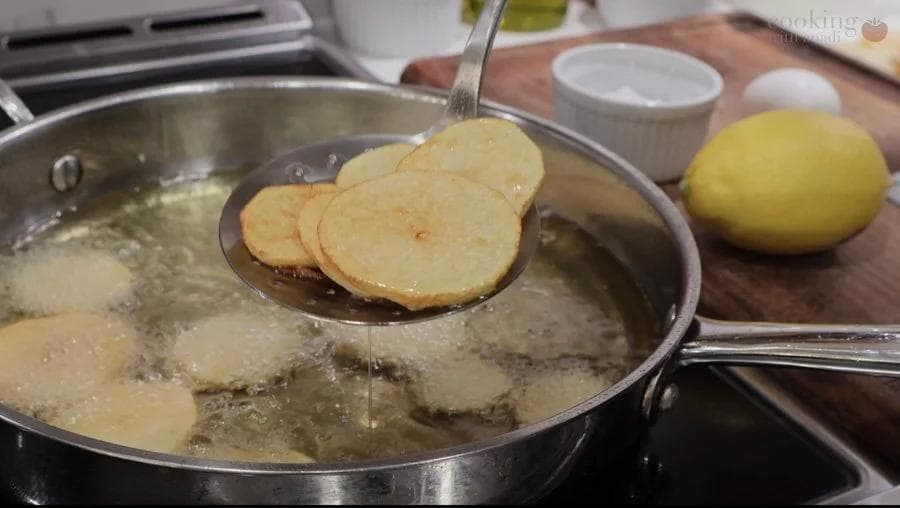 Sliced potatoes being shallow fried in oil in a stainless steel pan with a slotted spoon lifting golden-brown rounds.