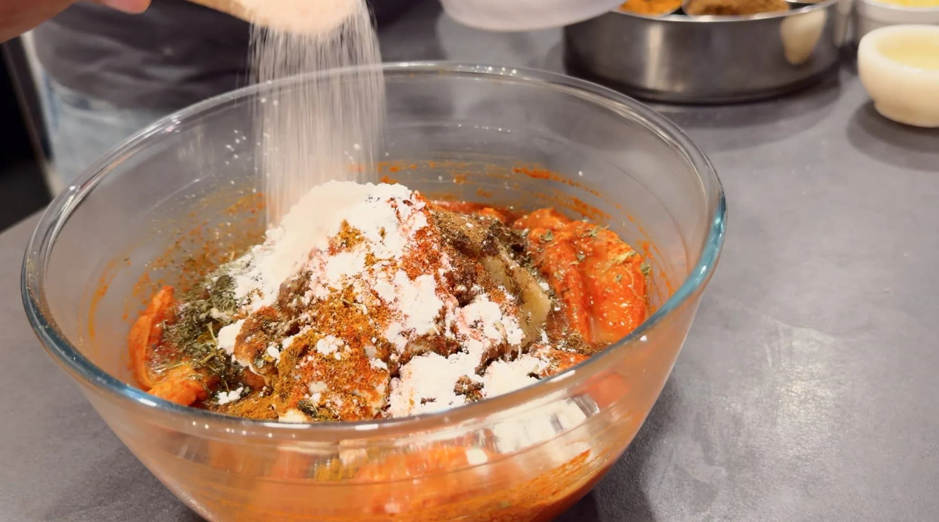 White powder being sprinkled over spiced chicken pieces with herbs and red marinade in a glass bowl.