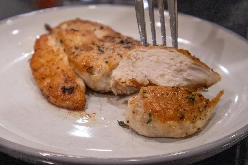 Pan-fried chicken breasts with golden-brown crust, seasoned with garlic and thyme, plated on white dish with fork.