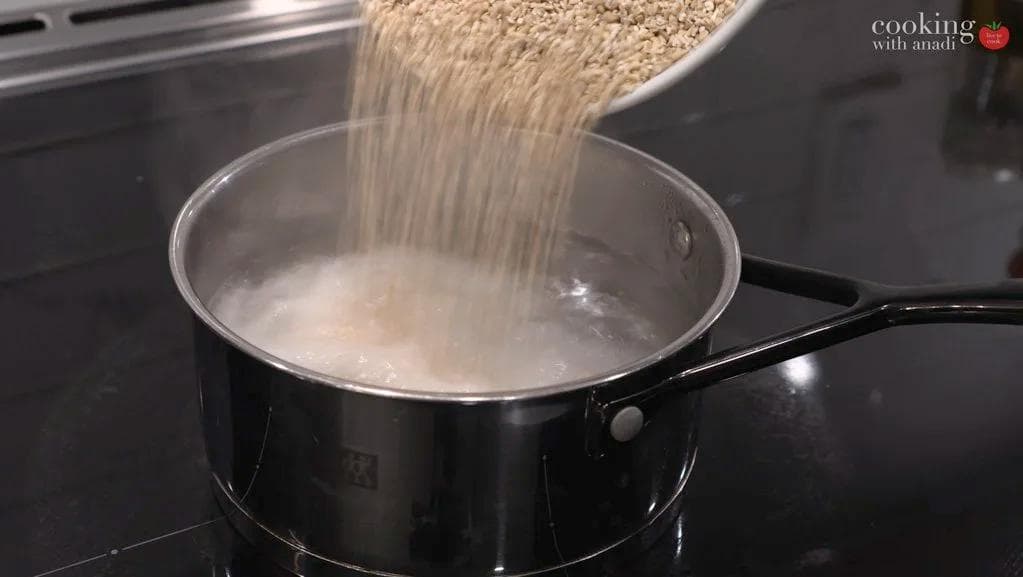 Steel-cut oats being poured into boiling water in a black saucepan on the stovetop.