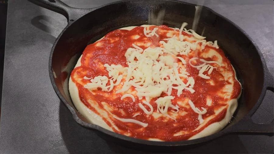 Cast iron skillet with pizza dough stretched to edges, topped with red sauce and shredded mozzarella cheese.
