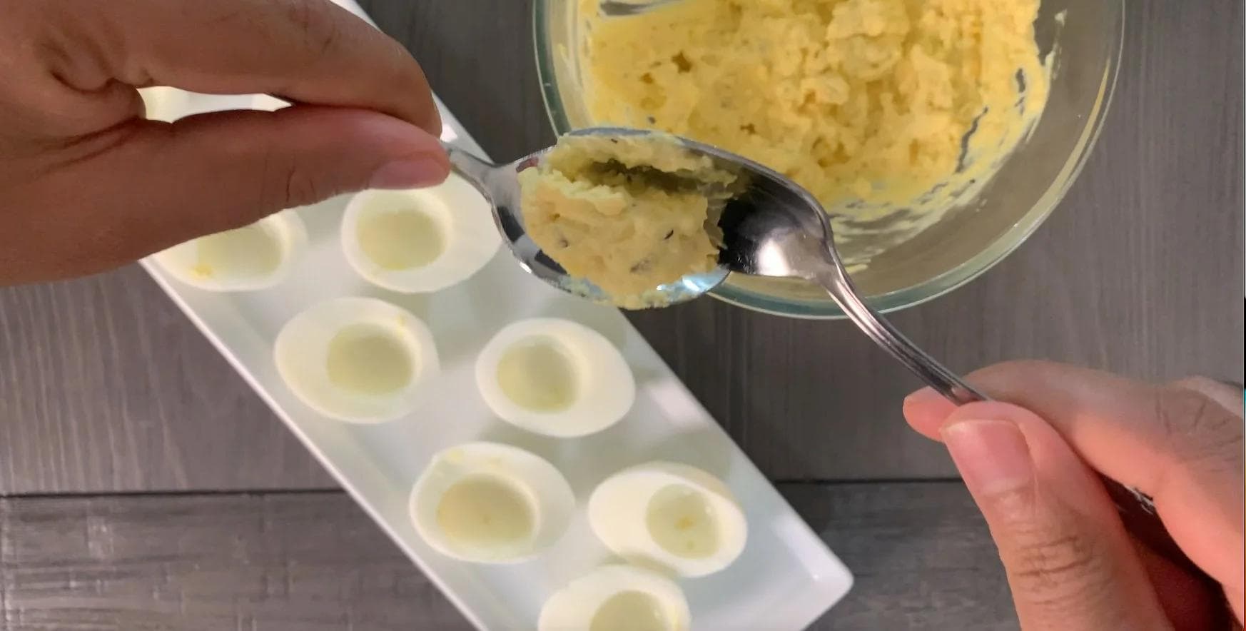Scooping deviled egg filling into egg white halves using a spoon from a bowl of creamy yolk mixture.
