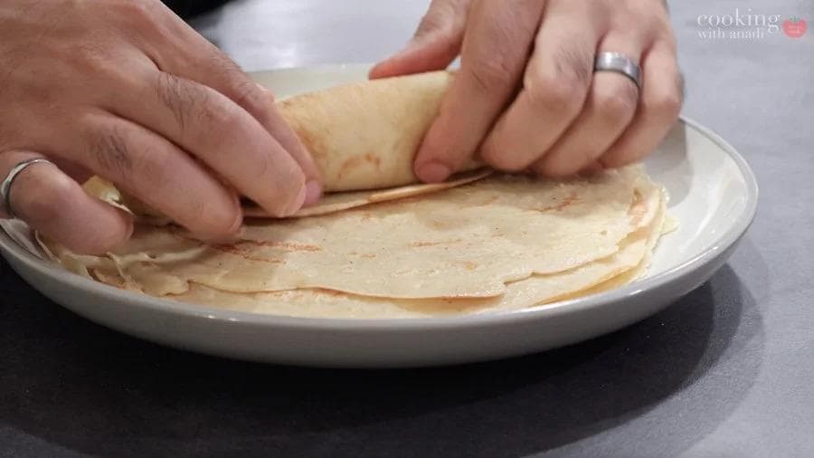 Hands rolling a pumpkin crepe with filling into a cigar shape on a white plate.