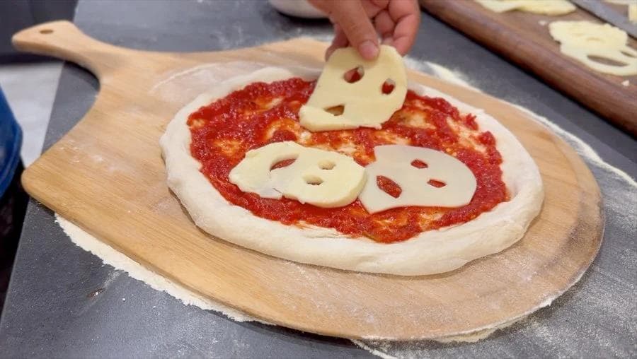 Ghost pizza with mozzarella cheese cutouts arranged on tomato sauce, placed on a wooden pizza peel.