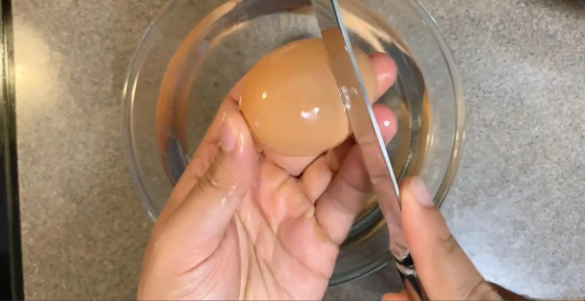 Hands peeling a boiled egg under water in a glass bowl, removing shell pieces.