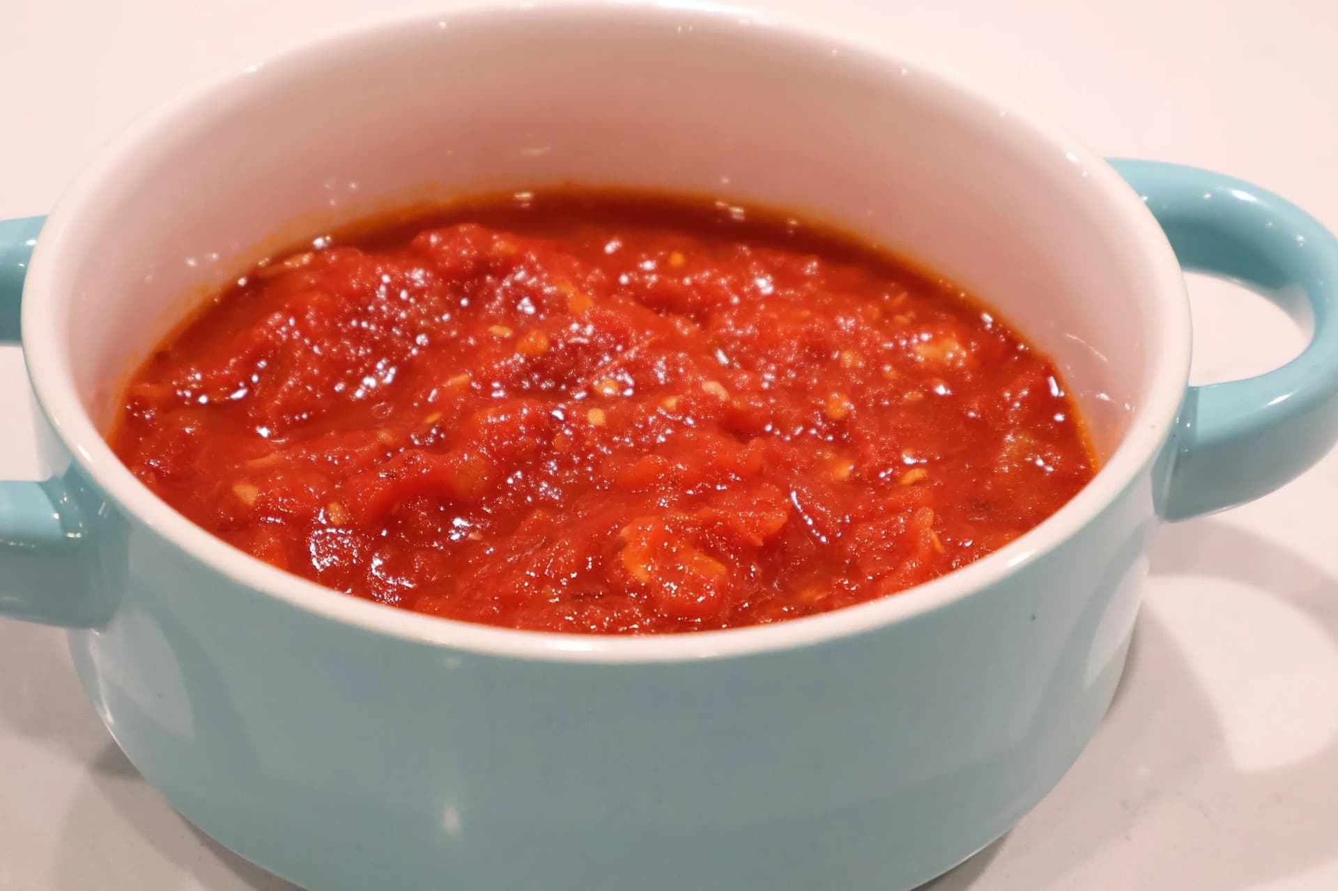 Chunky homemade marinara sauce in a light blue ceramic pot with handles, filled with bright red crushed tomatoes.