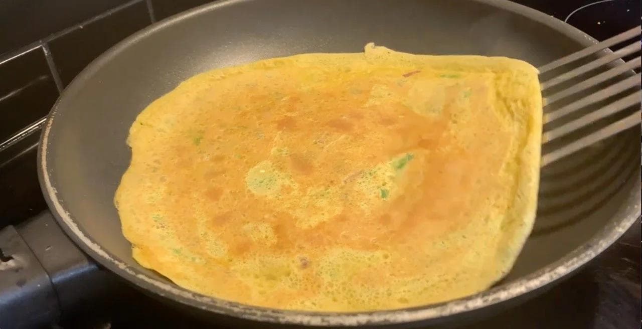 Golden-brown besan chilla cooking in a pan with crispy edges beginning to separate from the sides.