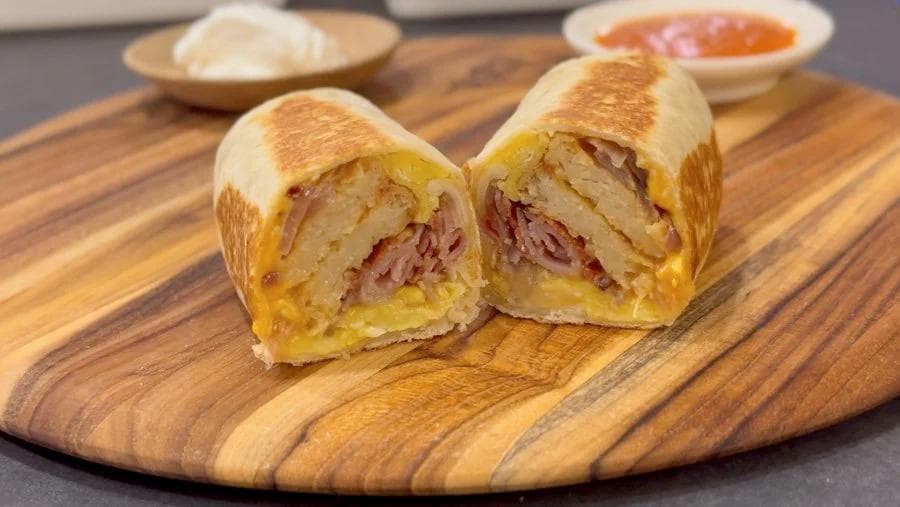 Two breakfast burritos cut in half on a wooden board, showing ham and egg filling inside toasted tortillas.