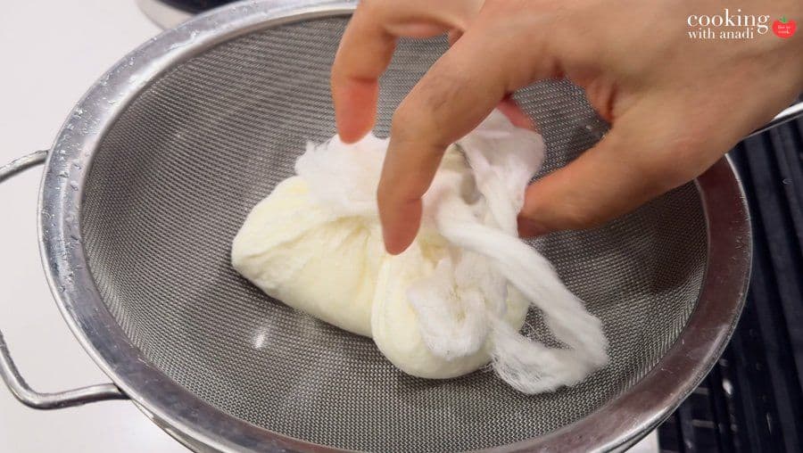 Hands gathering cheesecloth around drained hung curd in a metal sieve strainer.