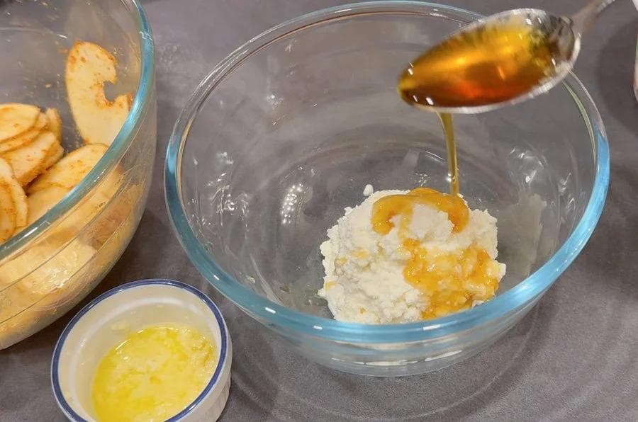 Maple syrup being drizzled into a bowl of ricotta mixture next to melted garlic butter and sliced potatoes.