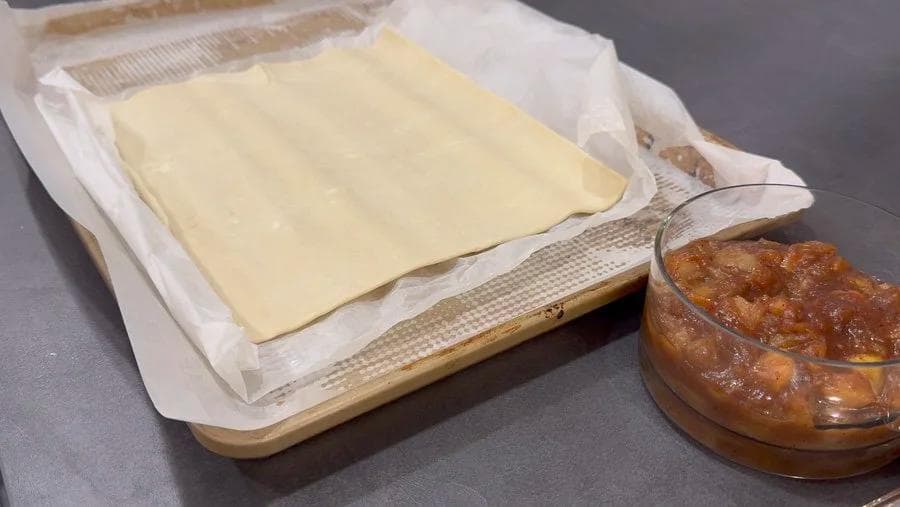Thawed puff pastry sheet on parchment paper with apple pie filling in a bowl nearby, ready for assembly.