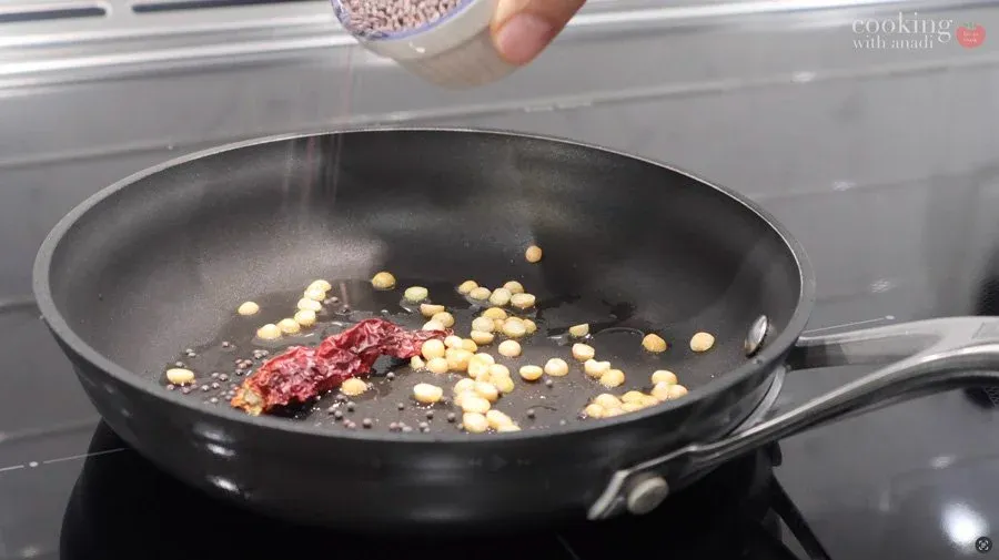 Dried red chilies, chana dal, and mustard seeds being toasted in coconut oil in a black pan on a stovetop.