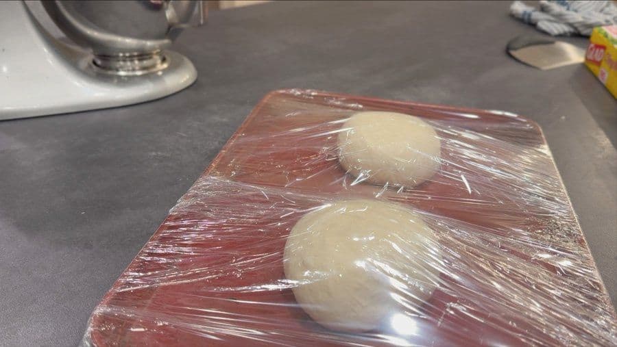 Two pizza dough balls wrapped in plastic wrap on a red baking sheet after cold fermentation.