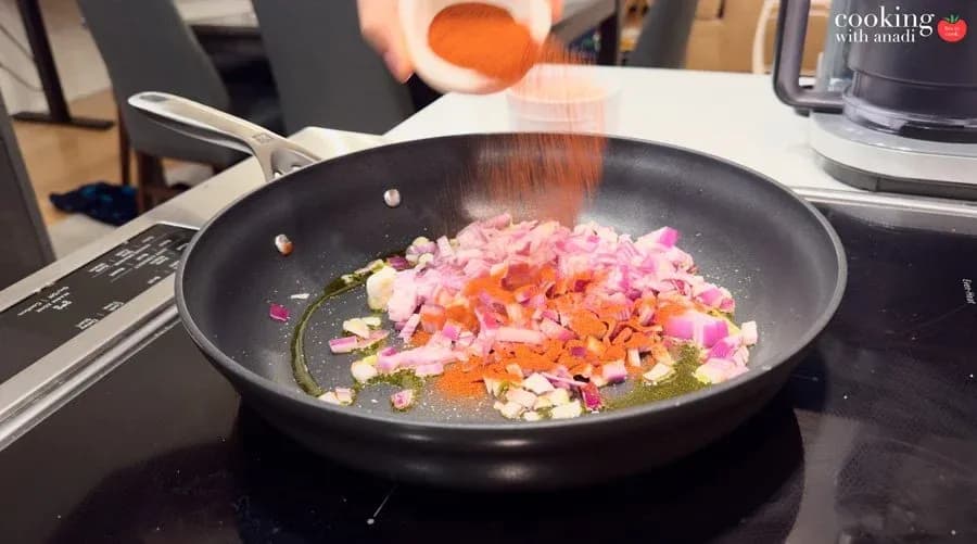 Red spice being sprinkled over diced red onions and carrots sautéing in oil in a black nonstick pan.
