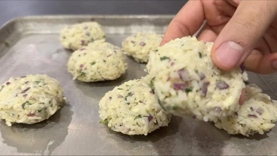 Uncooked Venezuelan rice fritters with visible cheese and herbs on a baking tray, being held up by a hand.