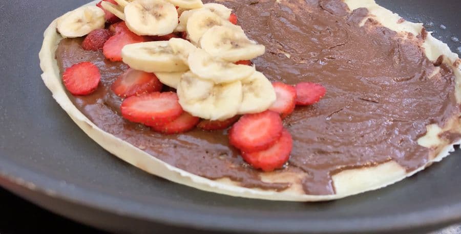 Nutella crepe topped with sliced strawberries and banana slices cooking in a gray pan.