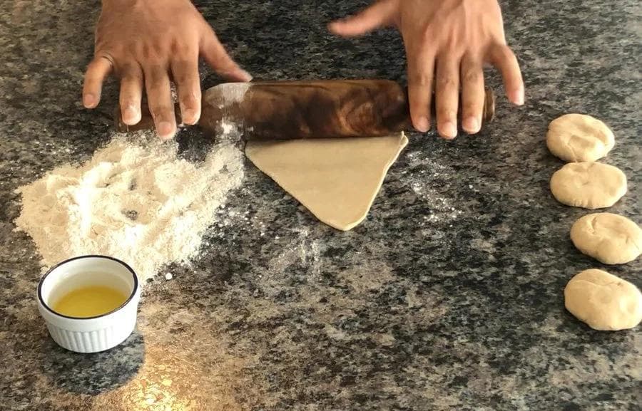 Hands rolling triangular paratha dough with rolling pin, ghee in bowl, and finished parathas on granite countertop.
