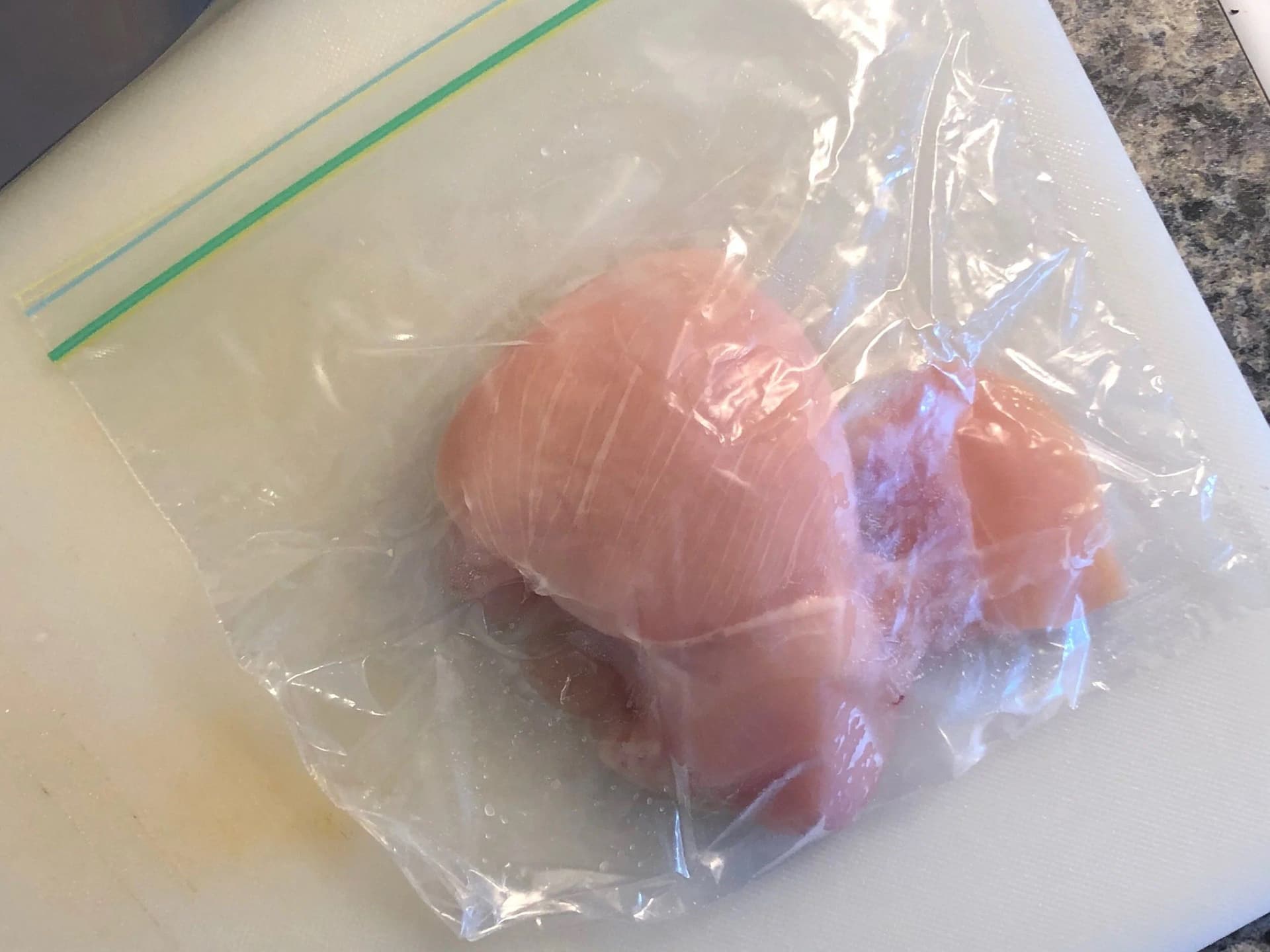 Raw chicken breast being pounded thin in a plastic bag with a meat tenderizer.