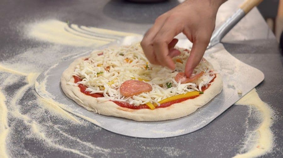 Hand placing pepperoni and shredded mozzarella cheese on pizza dough with tomato sauce and bell peppers.