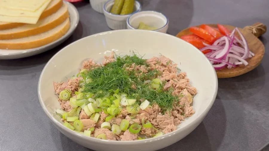 Tuna salad in a white bowl with diced green onions, fresh dill, and diced pickles, with bread and toppings visible nearby.