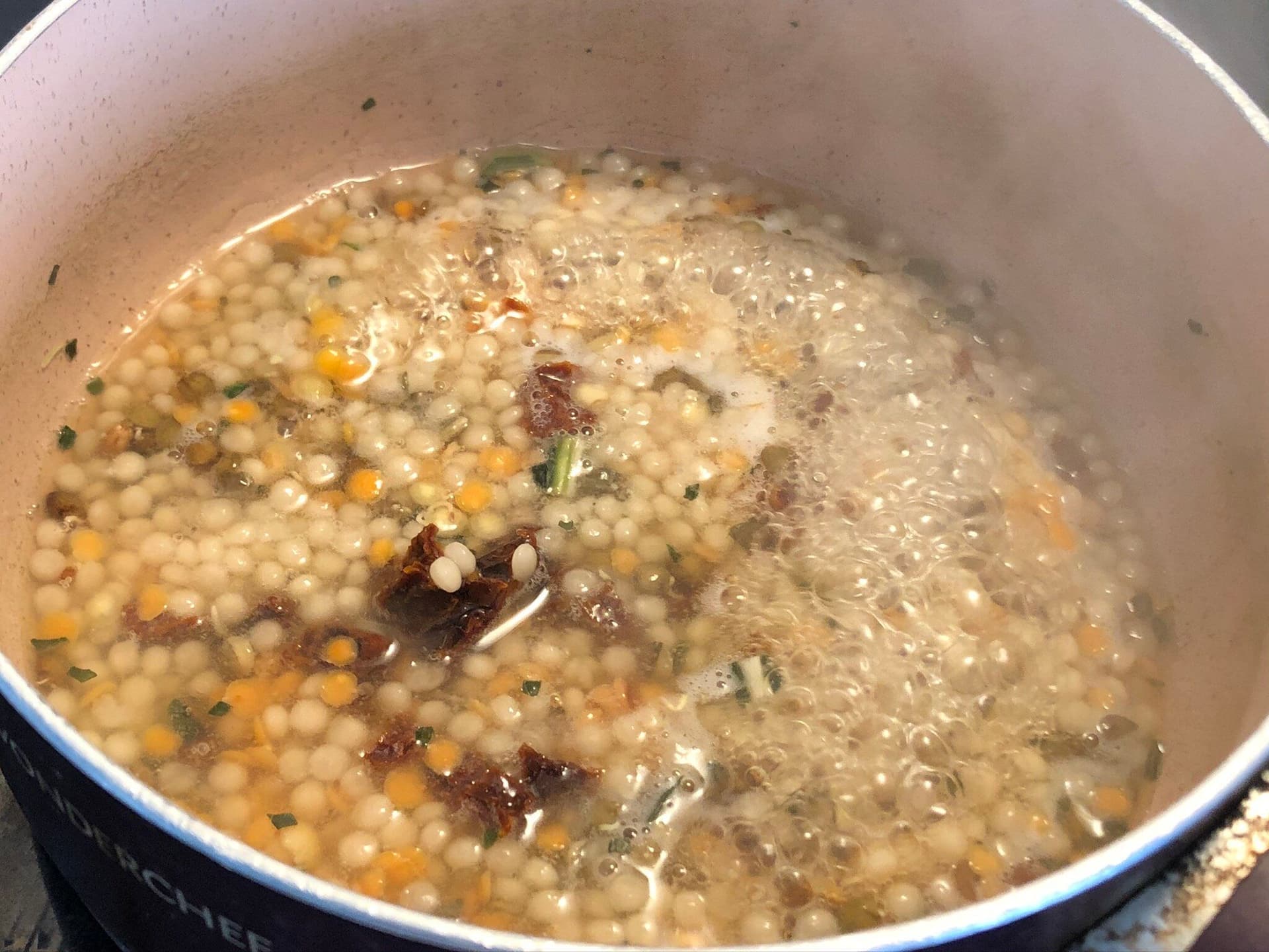 Couscous and lentil blend simmering in broth with sun-dried tomatoes and herbs in a pot.