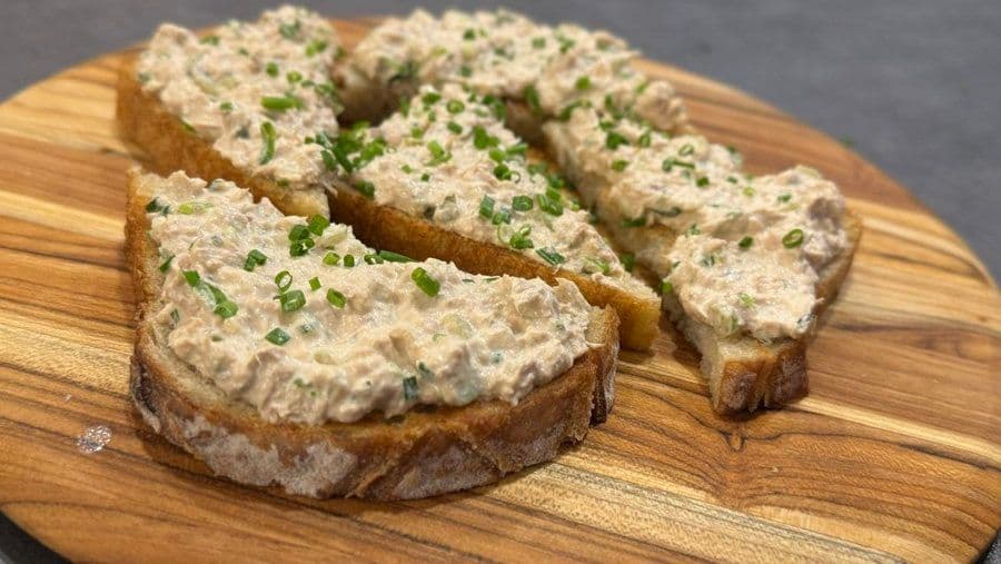 Tuna salad spread on toasted bread slices, garnished with chopped green onions on a wooden board.