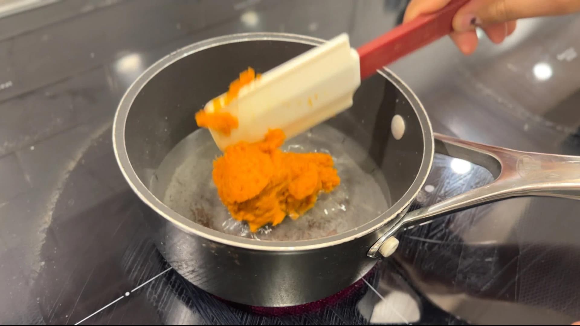 Orange pumpkin puree being added to a saucepan of clear sugar syrup with a spatula.