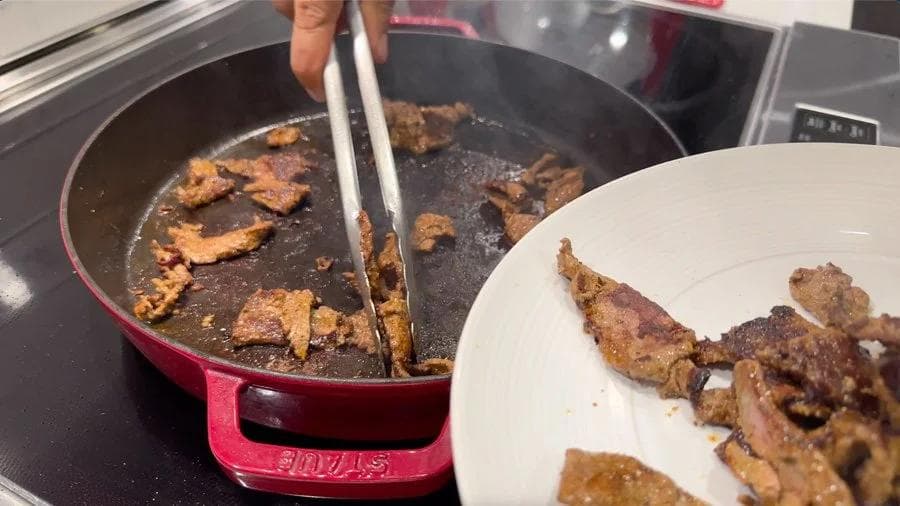 Browned steak strips being removed from a red cast-iron pan with tongs, with cooked meat on a white plate nearby.