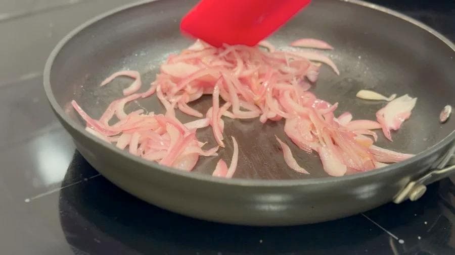 Sliced pink onions being sautéed in a pan with a red spatula, cooking step for breakfast burritos.