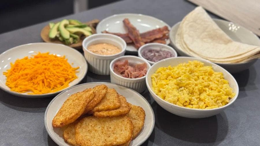 Breakfast burrito ingredients laid out including tortillas, scrambled eggs, shredded cheese, bacon, hash browns, and condiments.