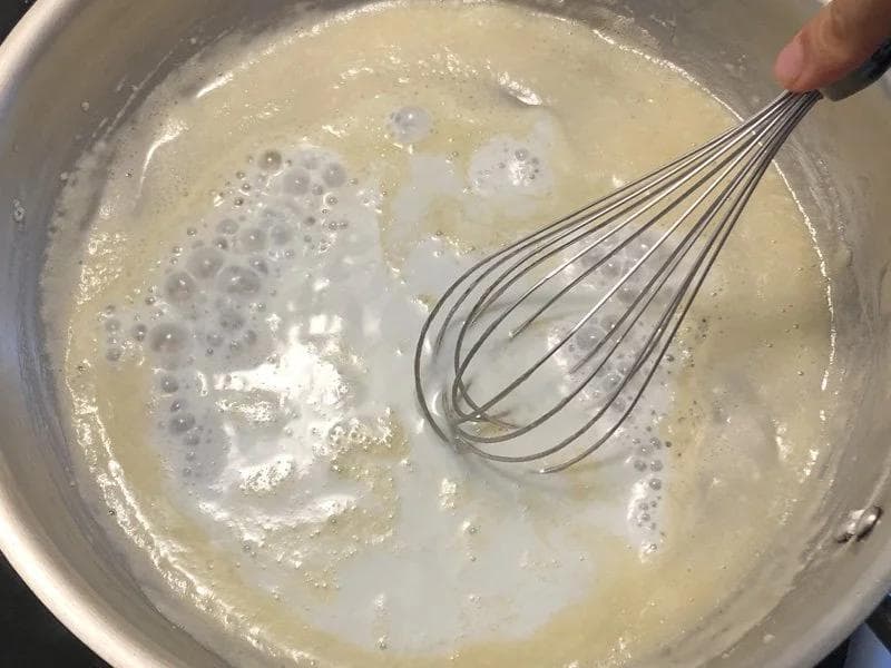 Whisking milk into a butter and flour roux to make béchamel sauce in a stainless steel pot.