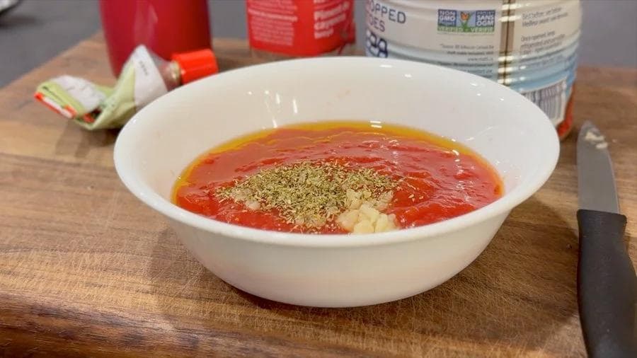 Pizza sauce mixed in white bowl with tomato base, minced garlic, and dried oregano sprinkled on top.