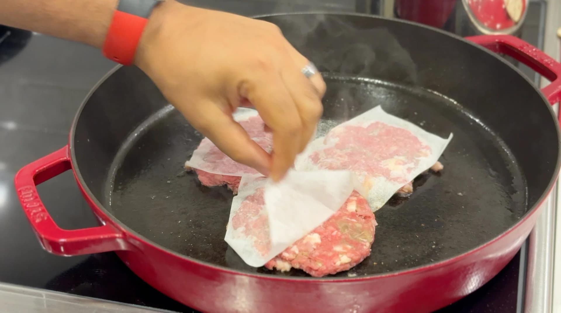 Hand placing raw burger patties on parchment paper into a red cast iron skillet for cooking.