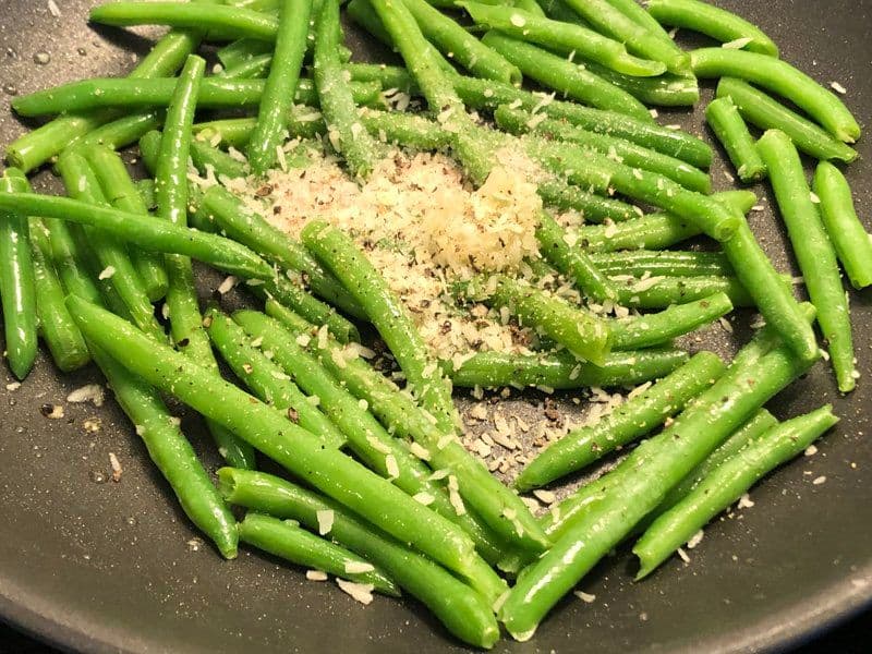 Green beans cooking in a dark pan with minced garlic and coconut flakes sprinkled on top.