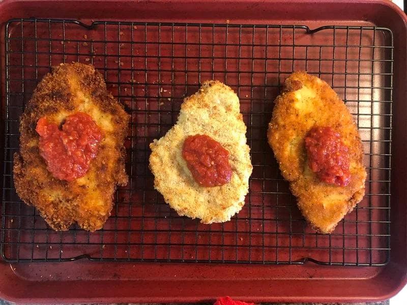 Three breaded chicken cutlets on a wire rack topped with marinara sauce, cooling after cooking.