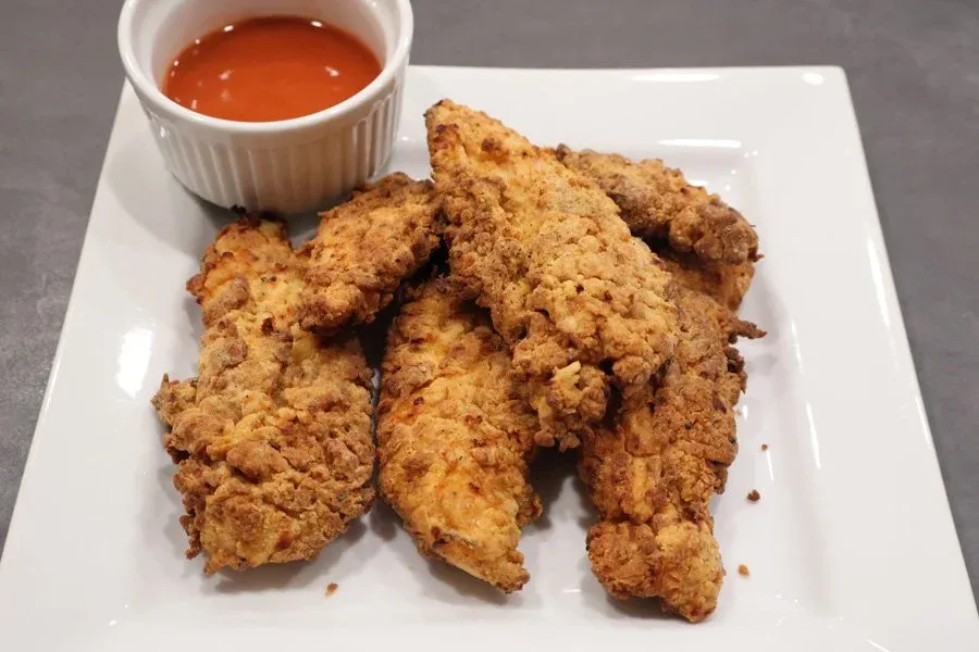 Crispy golden air fryer chicken tenders on a white plate with a small ramekin of red dipping sauce.