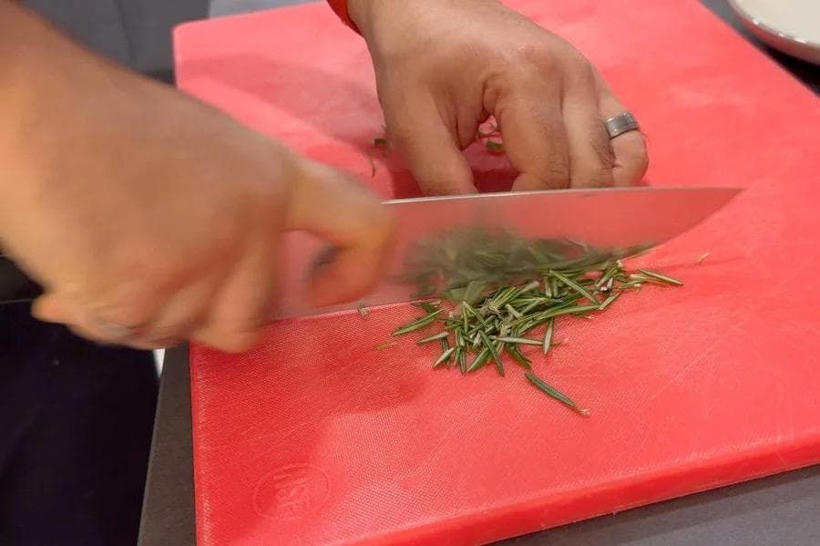 Hands chopping fresh rosemary with a knife on a red cutting board