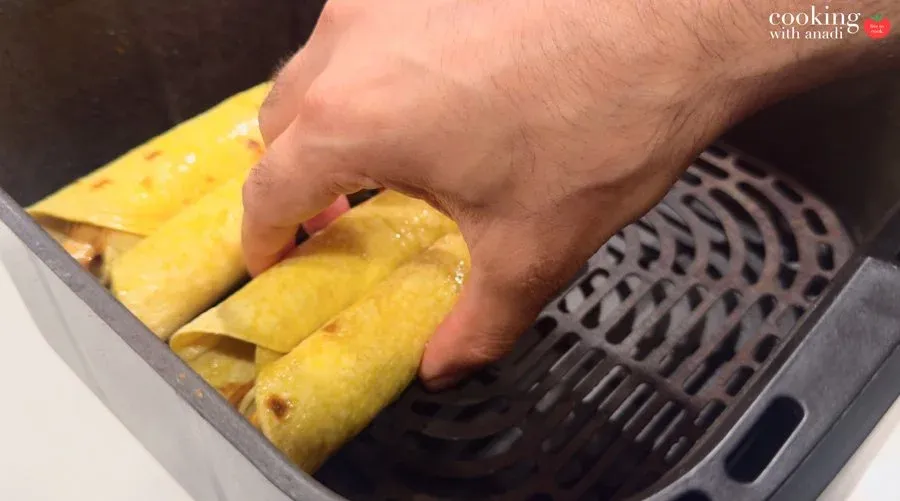 Hand placing rolled taquitos seam-side down into a black air fryer basket before cooking.