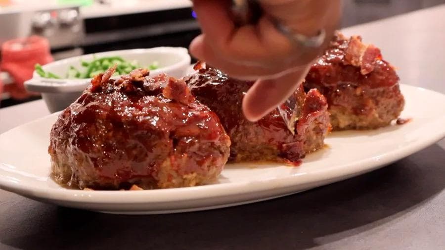 Three glazed chipotle mini meatloaves on a white plate, topped with bacon bits, with a side dish visible in background.