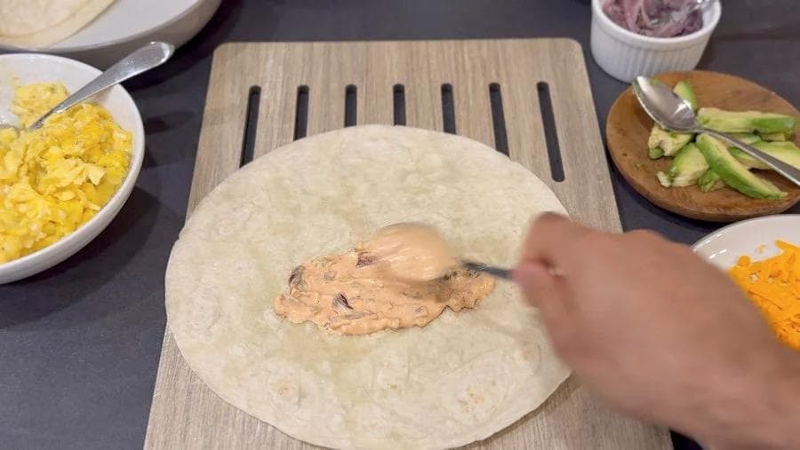 Hands wrapping shredded chicken and fillings in a flour tortilla on a wooden board with bowls of corn, avocado, and cheese nearby.