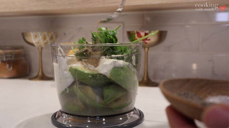 Food processor filled with avocado halves, lime, cilantro, and white creamy ingredients being blended together.