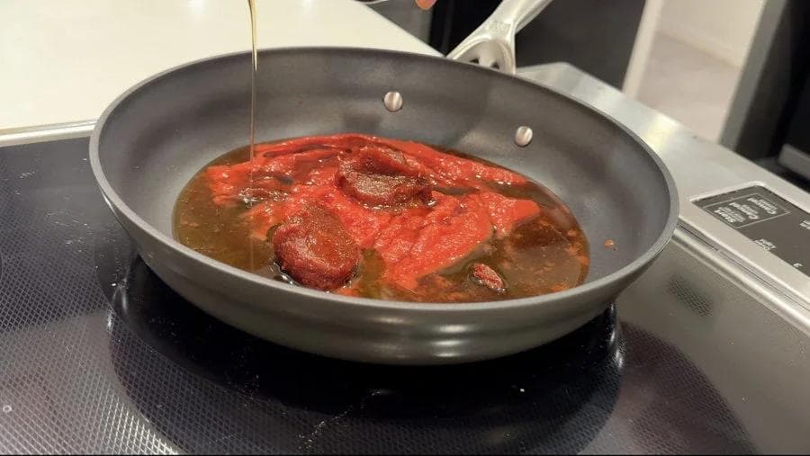 Red Korean sauce simmering in a gray pan on stovetop with oil being drizzled in.