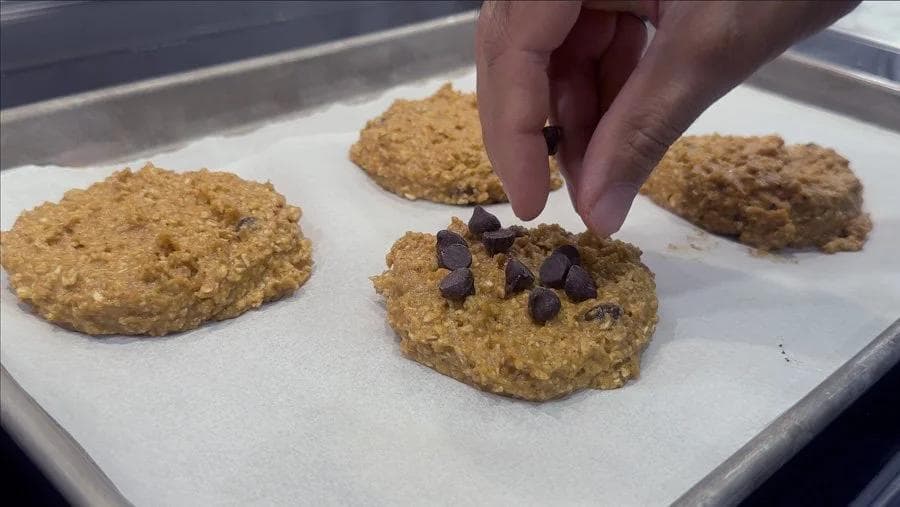 Hand placing chocolate chips on top of pumpkin cookie dough portions on parchment-lined baking tray.