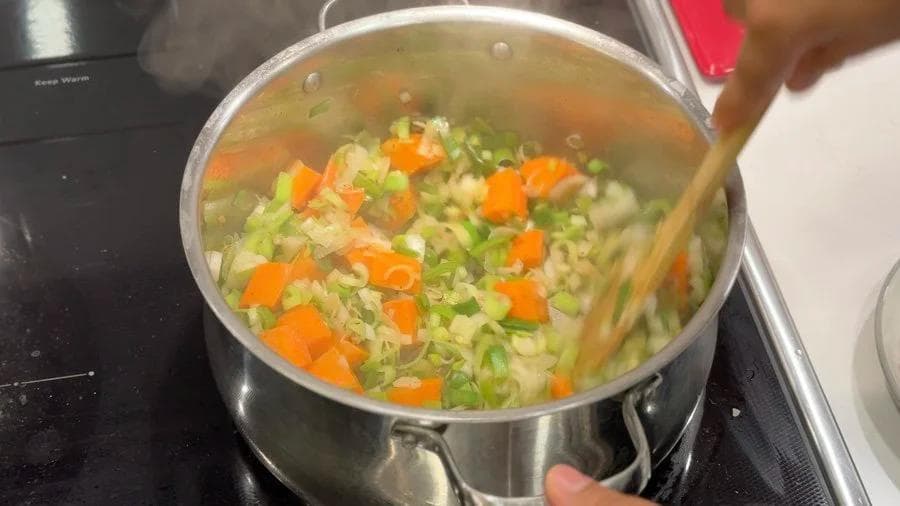 Sautéed vegetables including diced carrots, peas, and leeks in broth in a stainless steel pot on the stove.