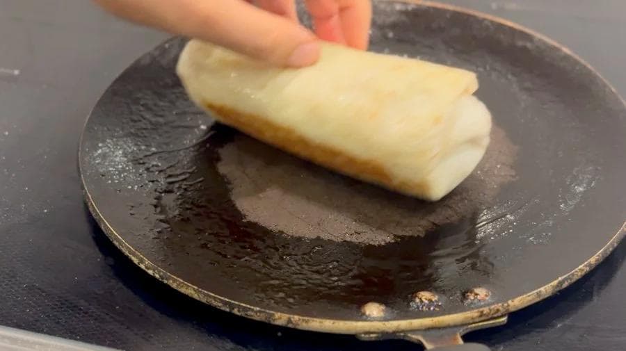Frozen breakfast burrito being placed on a dark skillet with melted butter for toasting.