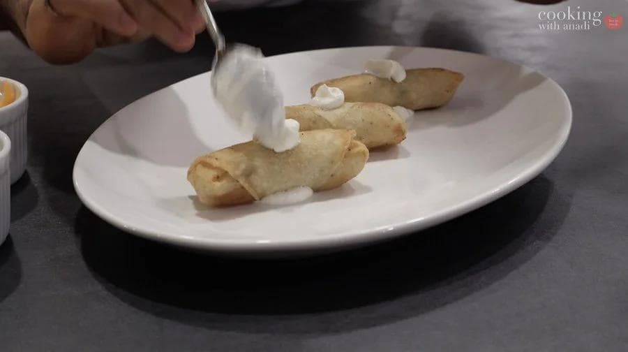 Crispy mashed potato spring rolls arranged on a white plate with dollops of sour cream being drizzled on top.