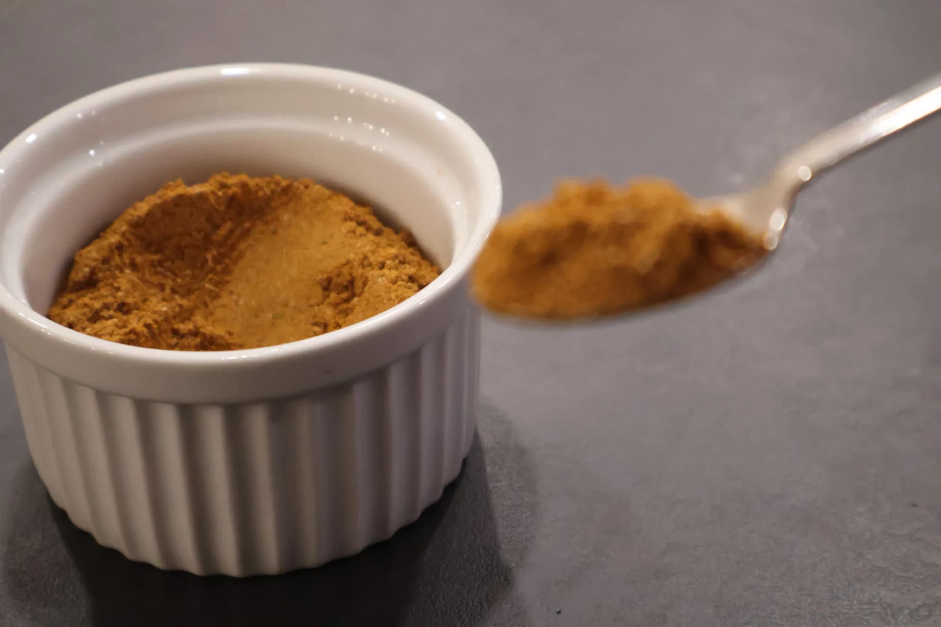 Nigerian curry powder in white bowl with spoon holding sample against gray surface